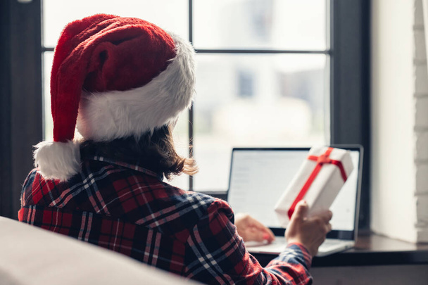 Woman in a red santa claus hat using laptop for making order sitting near window. Christmas online gifts shopping. Concept - Фото, зображення