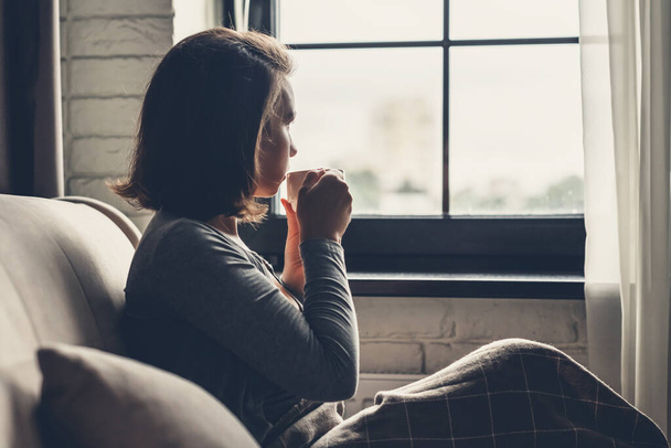 Επιστροφή άποψη της μοναχική νεαρή γυναίκα aucasian απολαμβάνοντας το πρωινό με φλιτζάνι ζεστό καφέ και κάθεται κοντά στο παράθυρο το πρωί.  - Φωτογραφία, εικόνα