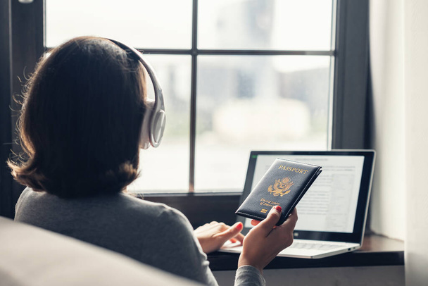 Woman holding american passport using laptop for making order and booking sitting near window in the headphones. Online service. concept. - 写真・画像