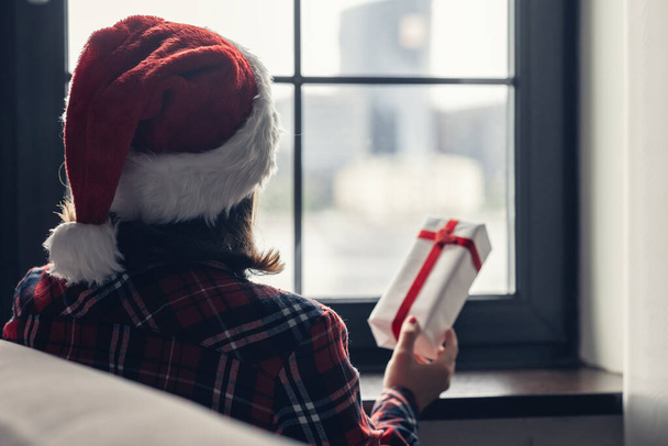 Back view of lonely sad young woman in a red santa claus christmas hat sitting near window and holding gift for boyfriend. Concept - Фото, зображення