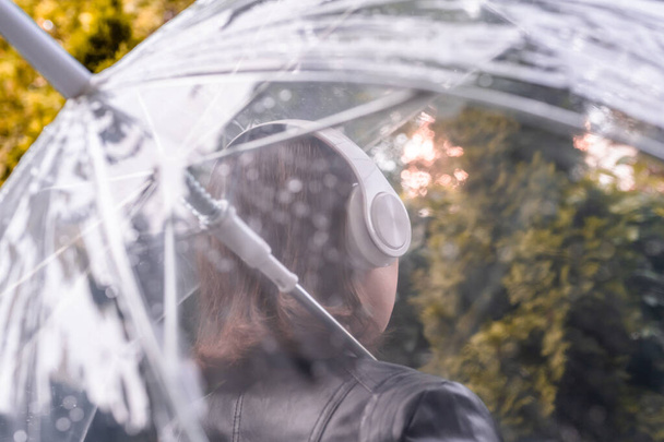 Autumn. Lonely sad woman in a headphones walking in a park, garden. View through wet  transparent umbrella with rain drops. Rainy day landscape.  - Foto, Bild