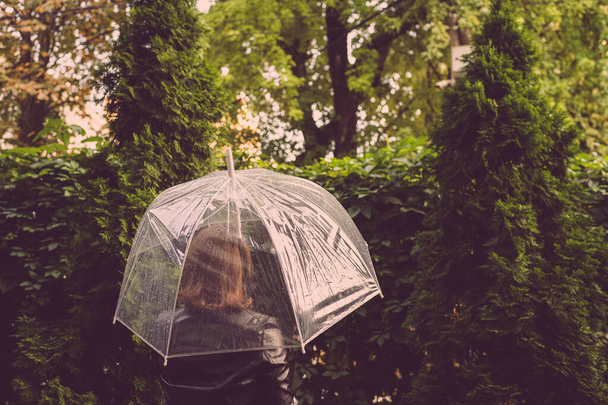 Autumn. Lonely redhead girl under a transparent umbrella with rain drops walking in a park, garden. Rainy day landscape - Photo, Image