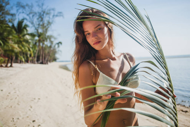 young sexy woman posing on sand beach under palm tree leaf, artistic shadow pattern on body, white bikini swim suit, tanned skin, slim body - Fotó, kép