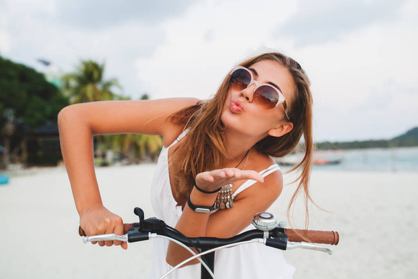 close up portrait of young attractive smiling woman in white dress riding on tropical beach on bicycle sunglasses traveling on summer vacation in Thailand, boho fashion style - Фото, изображение