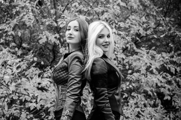 Two attractive women in autumn park wear leather jackets. Beautiful stylish city ladies on a walk. Concept of woman`s friendship and sister relationship - Photo, Image