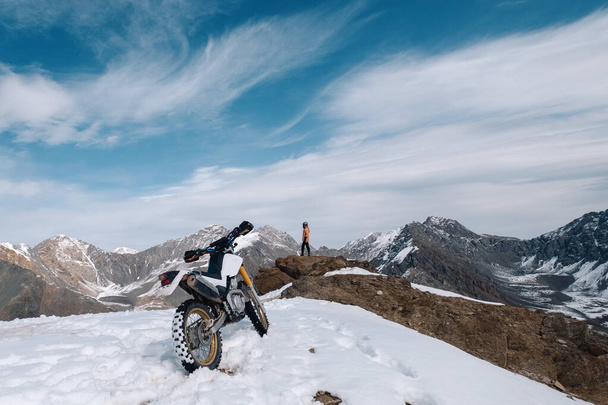 Female motorcyclist standing with her enduro motorcycle on snowy mountain top, snow peaks skyline view, high beautiful mountains - Foto, Bild