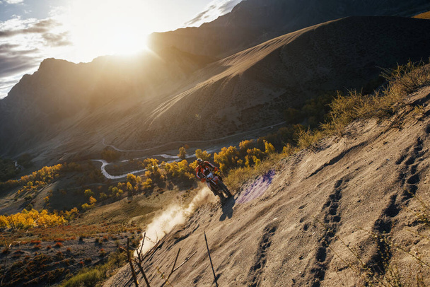 Extreme Rider escalando la cima de la montaña de arena en motocicleta enduro todoterreno. Hermoso paisaje montañoso en el fondo, colorido bosque otoñal y río bajo el sol  - Foto, imagen