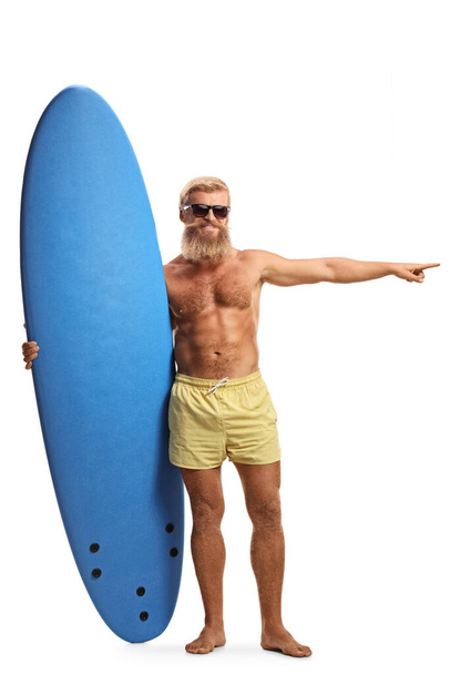 Full length portrait of a bearded surfer with a surfboard pointing to the side isolated on white background - Photo, Image
