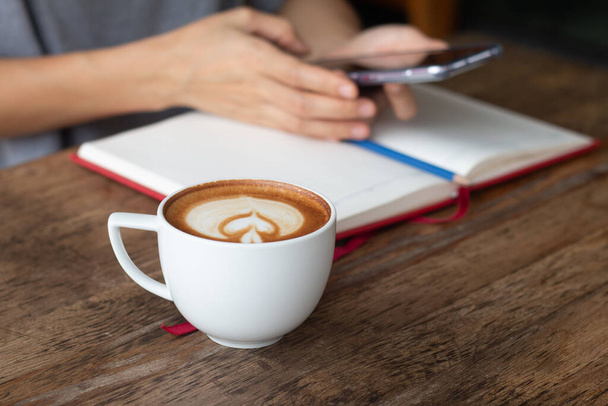 Frau, die ihre Arbeit auf dem Smartphone überprüft, verschwommen auf dem Hintergrund mit wärmendem Getränk, später Kaffee in einer Tasse vor sich auf dem Holztisch. - Foto, Bild