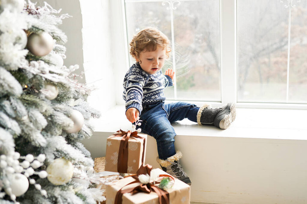 Noel ağacının yanındaki pencere eşiğinde oturan çocuk kahverengi hediyelerle, mutlu yıllar. - Fotoğraf, Görsel