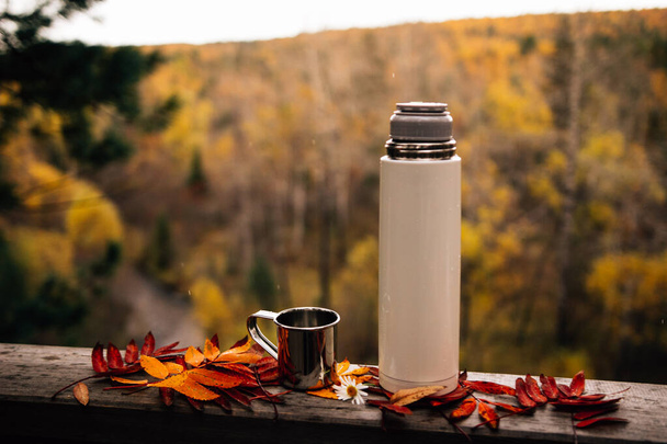Tasse mit Thermosflasche auf Holzzaun mit herbstlichem Laub  - Foto, Bild
