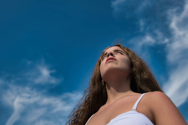 Tiener vrouw kijkend naar de horizon in een lage hoek schot op een blauwe hemel met wolken in de zomer. Overhead verlichting van de zon met veel detail op het gezicht van het model, haar en silhouet, kale schouders. - Foto, afbeelding