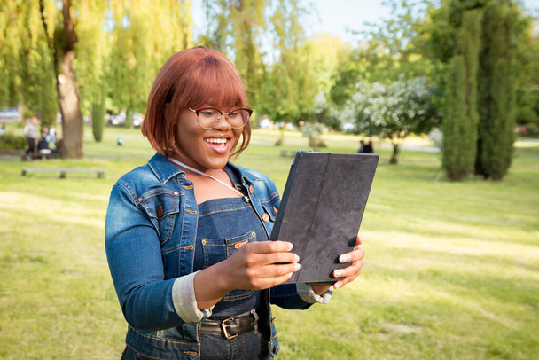 黒人の女子学生は現代的なコミュニケーション手段を使ってコミュニケーションをとる。屋外でタブレットを手にした女性の肖像画. - 写真・画像