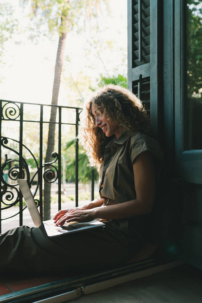Happy stylish female blogger smiling while chatting with friends in social networks on laptop device using wireless. Positive young woman shopping online while sitting on the balcony at home - Φωτογραφία, εικόνα