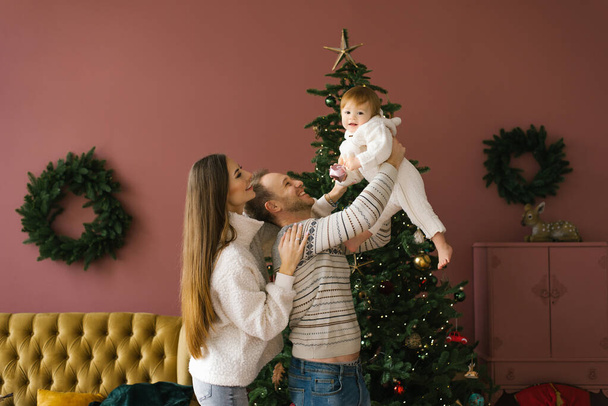 新しい家のリビングルームでクリスマスツリーの近くに小さな子供を持つスタイリッシュな若い家族の家族の肖像画。パパは娘を腕に乗せて - 写真・画像