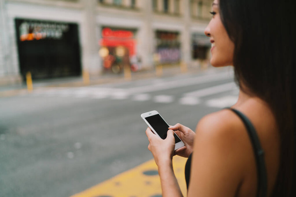 Cropped close up view of a mobile phone in the hands of a smiling brunette woman crossing the road. Photo with a focus on hipster girl's hands messaging on a smartphone on blurred street background - Foto, imagen