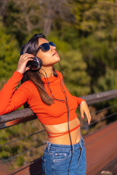 Lifestyle, brunette Caucasian girl enjoying music with headphones in a park. Young girl in red t-shirt and jeans, vertical photo - Foto, immagini