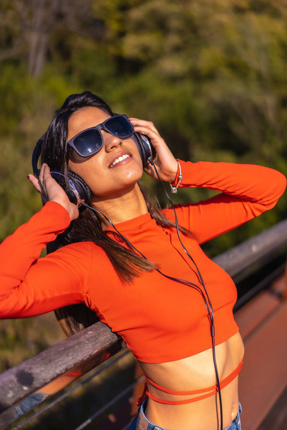 Lifestyle, brunette Caucasian girl enjoying music with headphones in a park. Young girl in red t-shirt and jeans - 写真・画像
