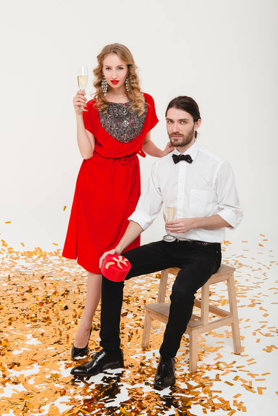 young stylish couple in love on white background holding glasses and drinking champagne, celebrating new year, wearing red dress, fashion jewelry, bow tie, happy disco party, having fun - Foto, Imagem