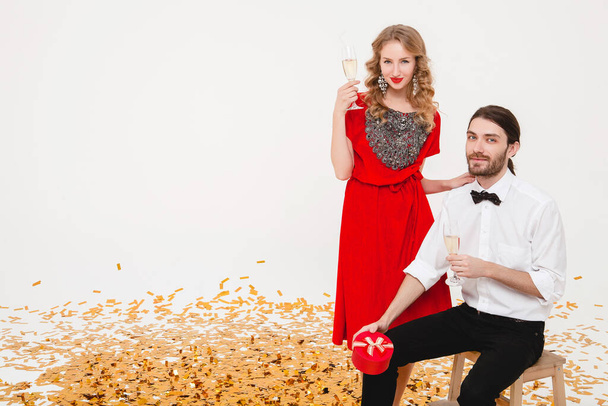 young stylish couple in love on white background holding glasses and drinking champagne, celebrating new year, wearing red dress, fashion jewelry, bow tie, happy disco party, having fun - Foto, Bild