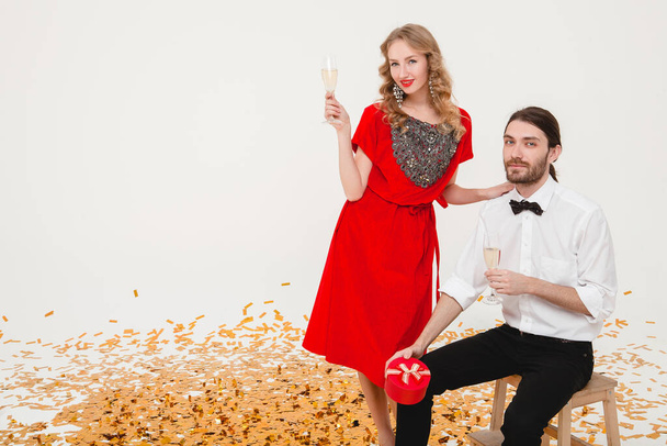 young stylish couple in love on white background holding glasses and drinking champagne, celebrating new year, wearing red dress, fashion jewelry, bow tie, happy disco party, having fun - Zdjęcie, obraz