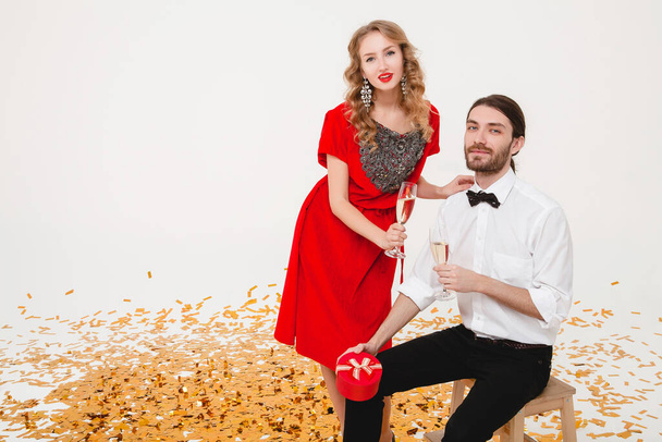 young stylish couple in love on white background holding glasses and drinking champagne, celebrating new year, wearing red dress, fashion jewelry, bow tie, happy disco party, having fun - Photo, image