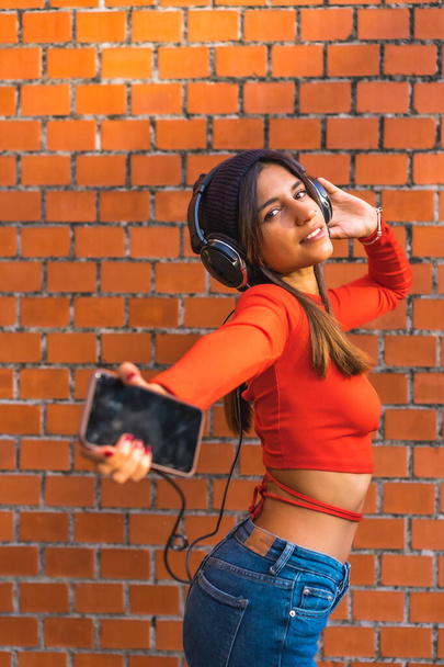 Lifestyle, young brunette with a wool cap and a red sweater enjoying music from her cell phone with her black headphones. Background of a brick wall. Vertical photo - Foto, Bild