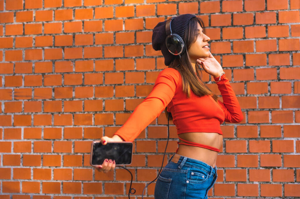 Lifestyle, young brunette with a wool cap and a red sweater enjoying music from her cell phone with her black headphones. Background of a brick wall - Φωτογραφία, εικόνα