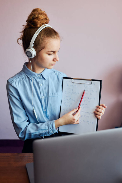 Young woman student showing homework, having classes, learning online, watching lesson remotely, listening to professor, talking with classmates on video call from home during quarantine. Young girl using laptop, headphones, books, manuals sitting at - Foto, Imagen