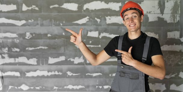 trabajador con casco naranja cerca de una pared de piedras. Los dedos índice de las manos indican un espacio vacío para el texto - Foto, imagen