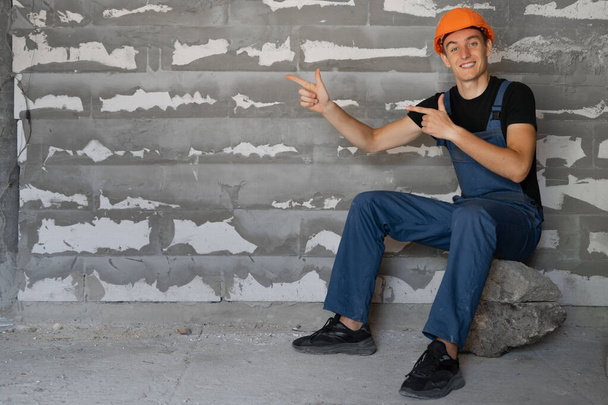 male builder dressed in work clothes and an orange mask. Sit on a stone with index fingers on an empty space for text. - Photo, Image
