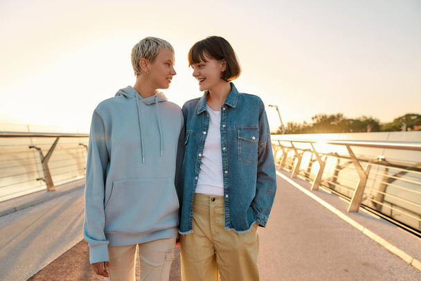 Köprüde yürürken ve birlikte güneşin doğuşunu izlerken mutlu lezbiyen çift birbirine bakıyor. Homoseksüellik, LGBT ve aşk konsepti - Fotoğraf, Görsel