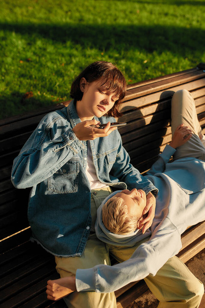 Young woman taking picture of her girlfriend while relaxing on the bench in the city park on a sunny day. Lesbian couple spending time together outdoors - Photo, Image