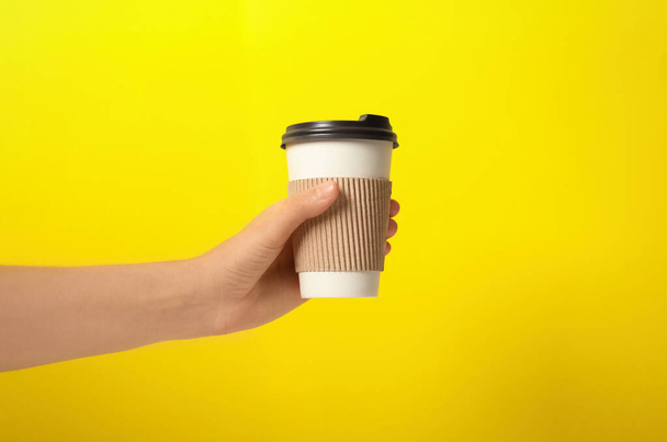 Woman holding takeaway paper coffee cup with cardboard sleeve on yellow background, closeup - 写真・画像