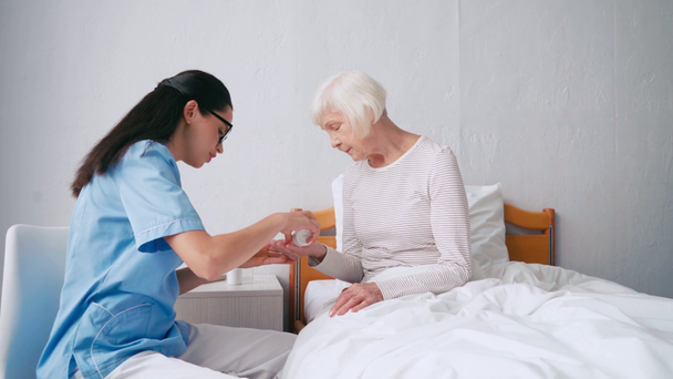 brunetka geriatrický zdravotní sestra dávat pilulky na stárnoucí žena sedí v posteli - Záběry, video