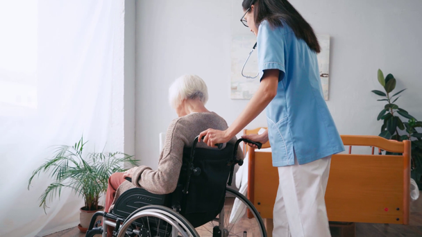brunette nurse moving aged woman in wheelchair around room - Záběry, video