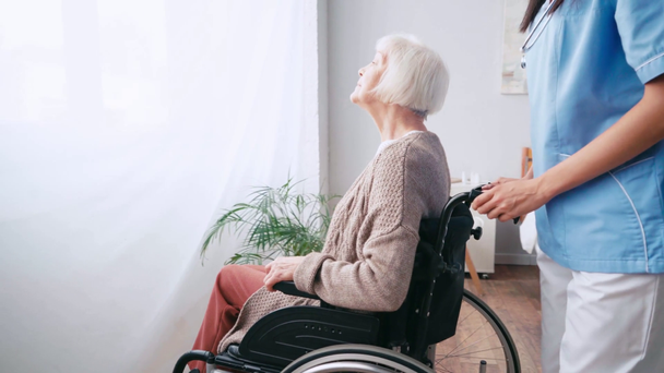 verpleegster verplaatsen glimlachende oudere vrouw in rolstoel naar raam - Video