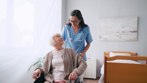 χαρούμενη νοσοκόμα κινείται χαμογελαστή ηλικιωμένη γυναίκα σε αναπηρική καρέκλα - Πλάνα, βίντεο