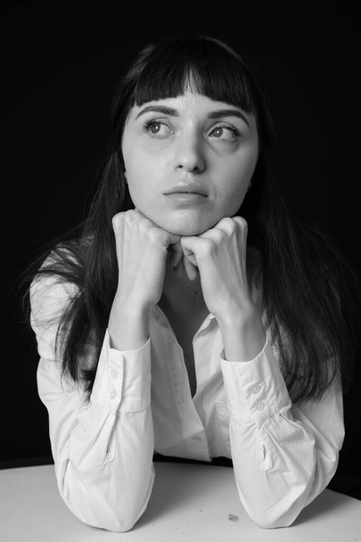 Студийный портрет красивой брюнетки в белой рубашке, сидящей за столом, на обычном черном фоне, смотрящей в сторону - Фото, изображение