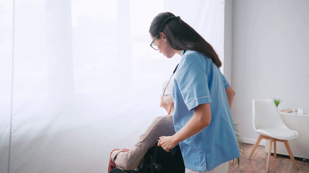 soeur gériatrique pointant avec la main tout en déplaçant le patient en fauteuil roulant à la fenêtre - Séquence, vidéo