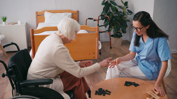 vrouw in rolstoel en verpleegster spelen rock-papier-schaar spel in de buurt van dominostenen - Video