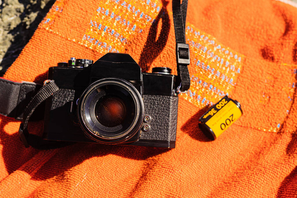 Antigua cámara de fotos analógica negra irreconocible sobre un fondo naranja con una película de carrete de 35 mm junto a ella. Encima de algunas rocas marinas en un día soleado en España. - Foto, Imagen