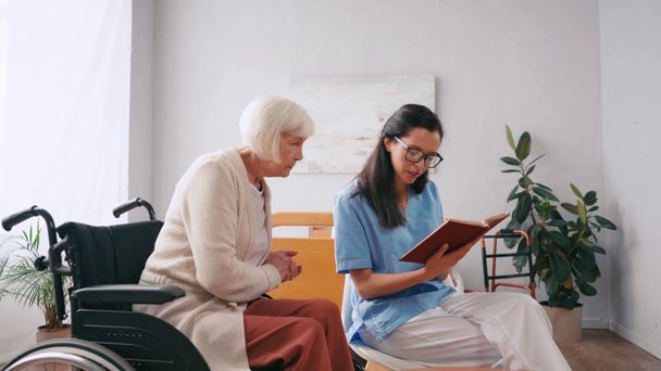 молодая брюнетка медсестра читает книгу пожилой женщине в доме престарелых - Кадры, видео