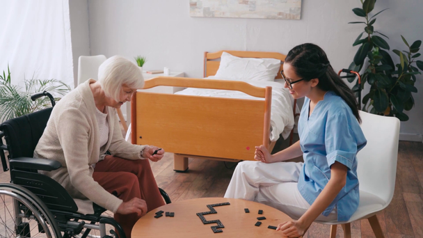 senior femme en fauteuil roulant jouer dominos avec jeune infirmière - Séquence, vidéo