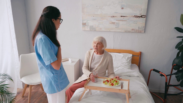 Esmer hemşire kahvaltıda yaşlı kadınla konuşurken el kol hareketi yapıyor. - Video, Çekim