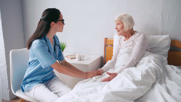 giovane geriatra e donna anziana che si tiene per mano e parla in clinica - Filmati, video