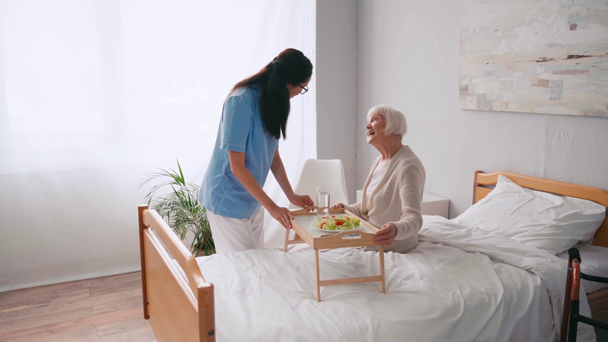 若い看護師がベッドで女性に朝食を持ってトレイを運ぶ - 映像、動画