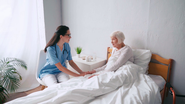 ηλικιωμένη γυναίκα που κάθεται στο κρεβάτι και μιλάει με μελαχρινή γηριατρική νοσοκόμα - Πλάνα, βίντεο
