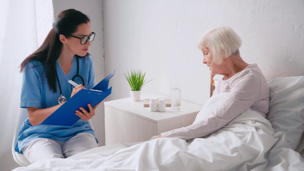 young geriatrician talking to aged woman and writing in clipboard - Footage, Video