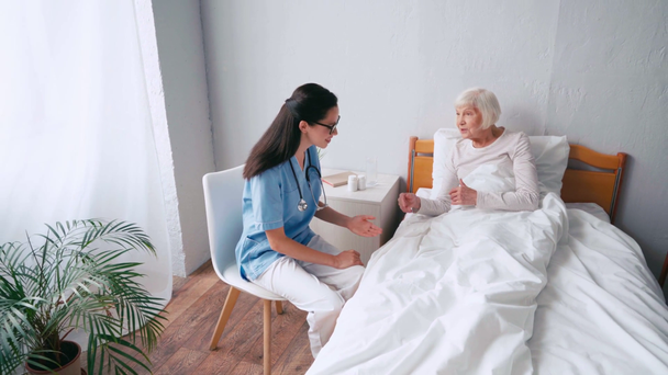 Yaşlı kadın yatakta uzanıp doktorla konuşurken el kol hareketi yapıyor. - Video, Çekim